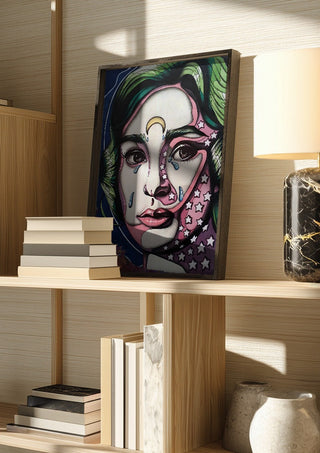 Audrey Hepburn by Okiana in a dark frame on a wooden shelf beside books and a lamp