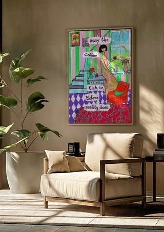 May the Coffee Kick In by Camilla Nilsson framed above a beige armchair beside a potted plant
