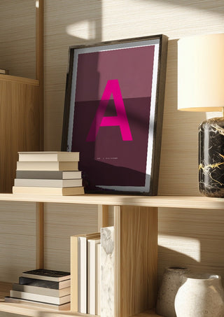 A as in Awesome – Cerise by Quite Nice Prints in a dark frame on a wooden shelf beside books and a lamp