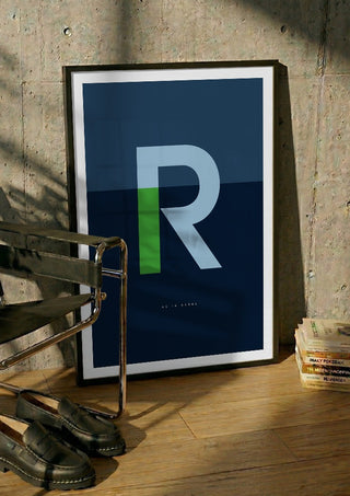 R as in Rebel – Blue by Quite Nice Prints in a black frame leaning against a concrete wall by a steel chair