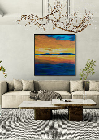 Under An Evening Sky by Finbar Stevens in a black frame above a beige sofa under a branch chandelier