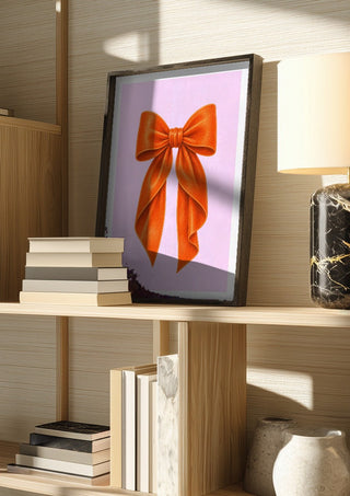 Orange velvet bow on pink by Ohkimiko in a dark frame on a wooden shelf beside books and a lamp