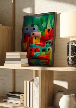 Houses of Favela by Nittaart in a dark frame on a wooden shelf beside books and a lamp