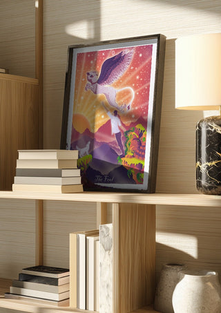 The Fool by Sciurus tarotus in a dark frame on a wooden shelf beside books and a lamp