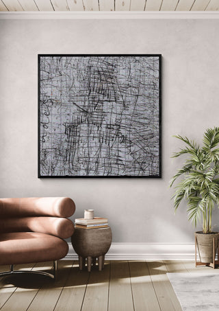 looks like a house by Priit Ruttas in a black frame on a pale wall above a tan leather chair and a potted palm