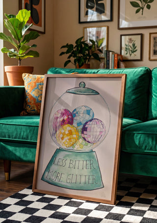 Less Bitter More Glitter by L.OE. Art in a wooden frame beside a green velvet sofa on a chequered floor