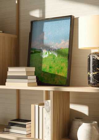 Touch Grass by Jakob Falkenberg in a dark frame on a wooden shelf beside books and a lamp